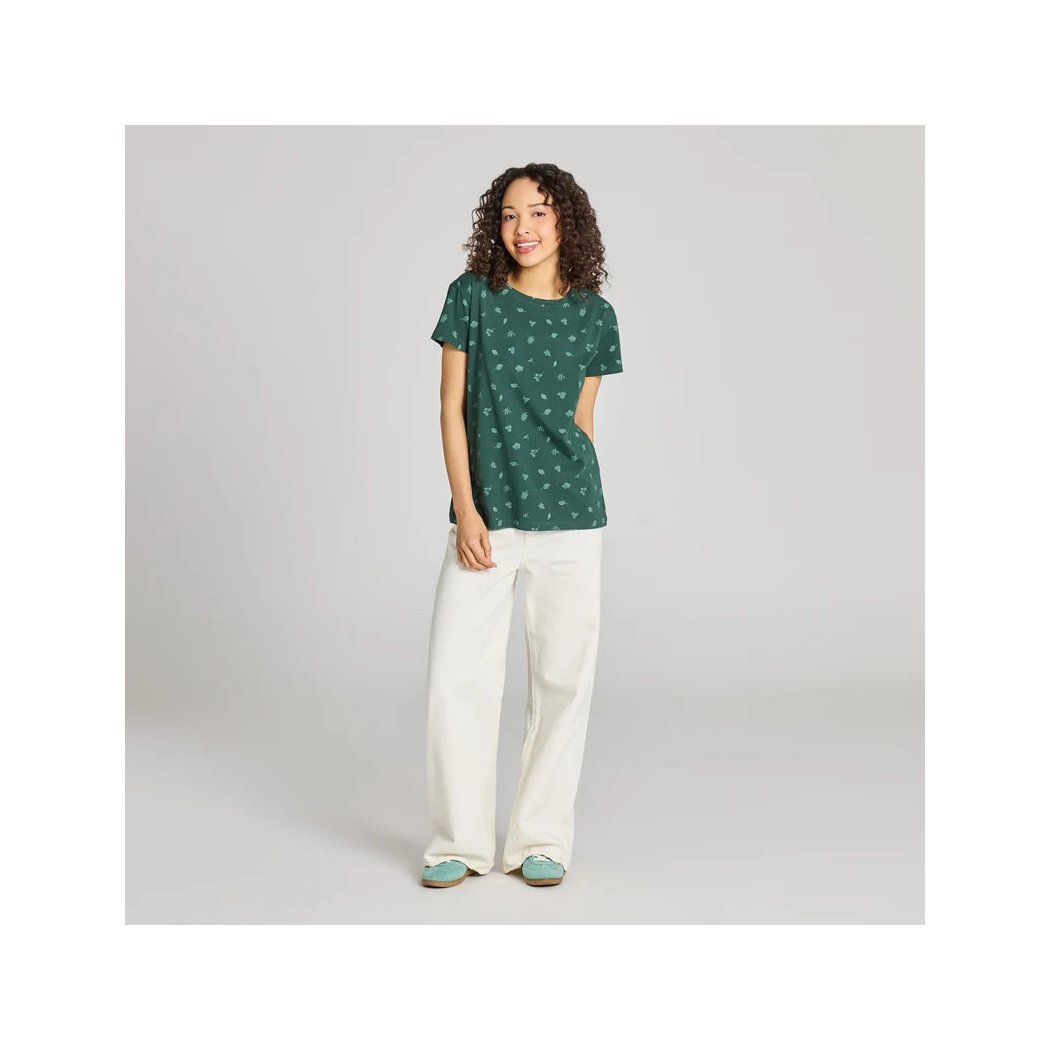 A model wears the Life is Good Women's Falling Leaves Pattern Crusher in Treeline Green with white pants against a gray background.