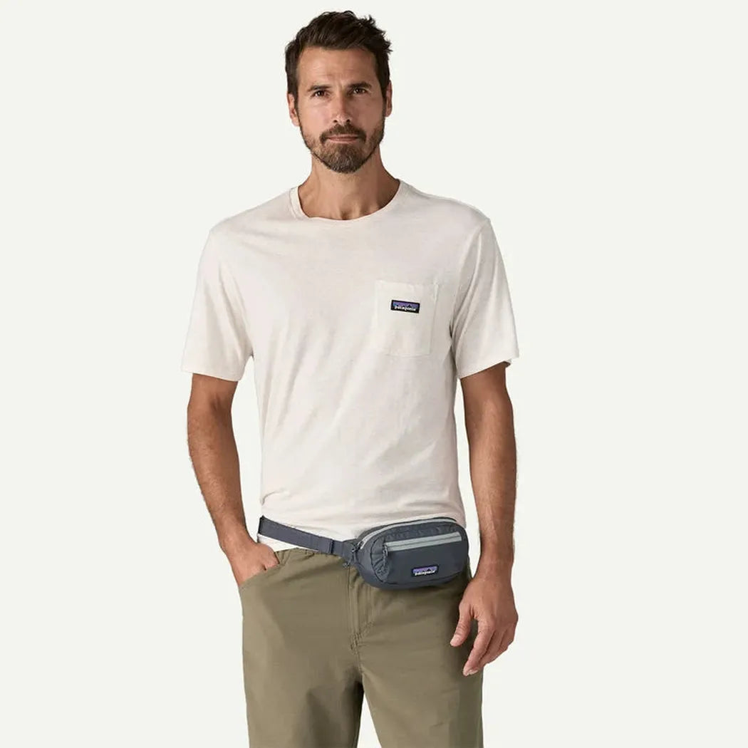 A man wearing a white Patagonia t - shirt and khaki pants stands against a white background, showing the Patagonia Terravia Mini Hip Pack in Aqua Stone around his waist.