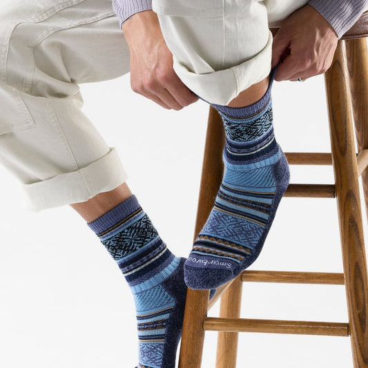 Model wearing Smartwool Everyday Cozy Fair Isle Sweater Crew Socks in Nightfall Blue, sitting on a wooden stool with rolled cuffs.