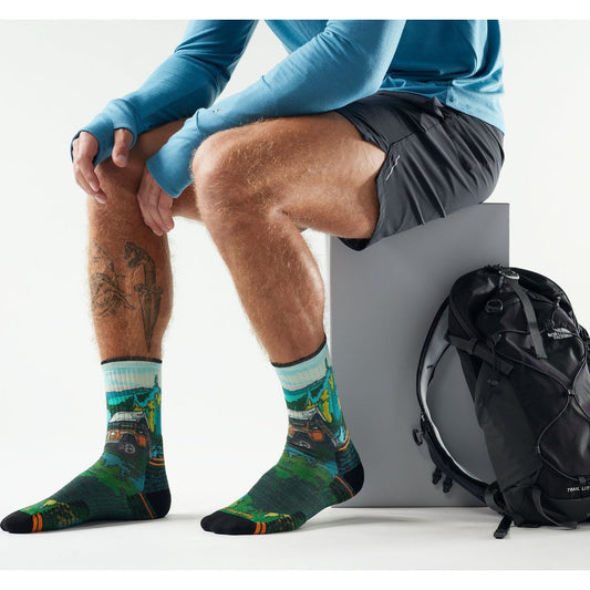Man wearing Winter Moss Smartwool Hike Trail Trekker Print Crew Socks sits on a gray block with a black backpack nearby.