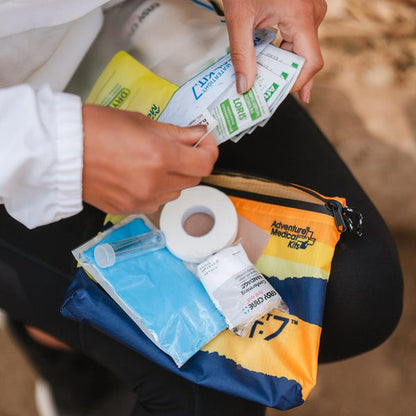 A person holds small packets over an Adventure Ready Brands AMK Ultralight/Watertight .7 first aid kit with a roll of tape and blue gauze visible.