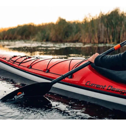 A person paddles a red Current Design kayak with a black Aqua Bound Sting Ray paddle in a calm waterway with trees in the background.