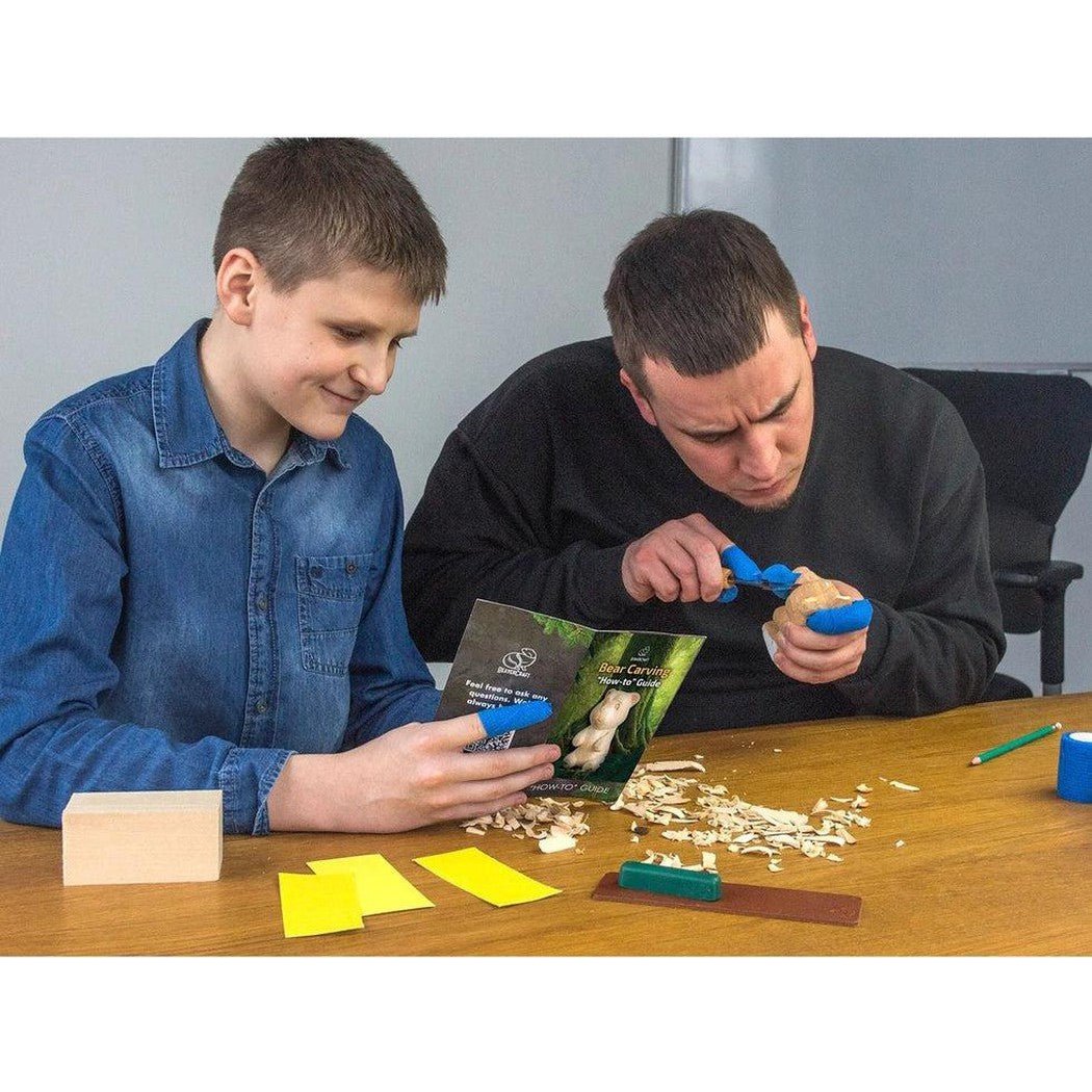 A man and boy work on a Beavercraft Bear Carving Kit at a table with wood shavings and carving tools.