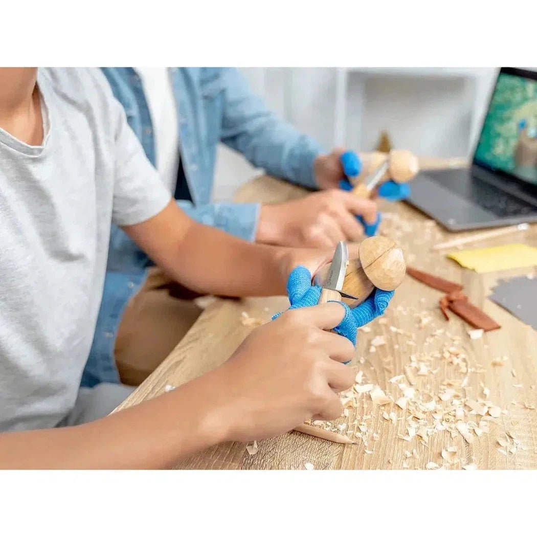 Two people use blue gloves with the Beavercraft Family Fun Wood Carving Kit on a wooden table with wood shavings and a laptop.