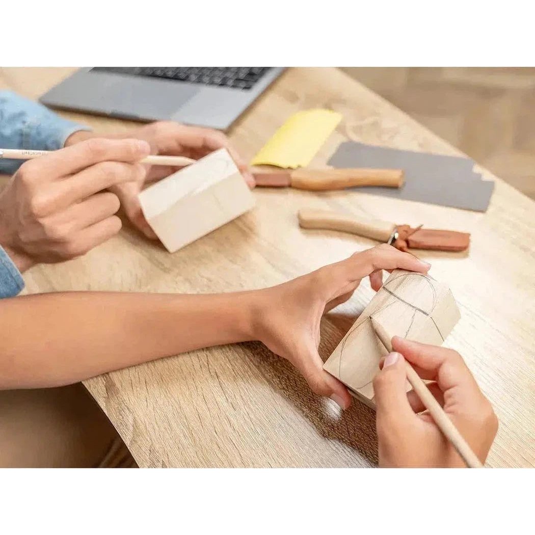Two people work on wood carving projects at a table with a laptop and tools in the background, using the Beavercraft Family Fun Wood Carving Kit.