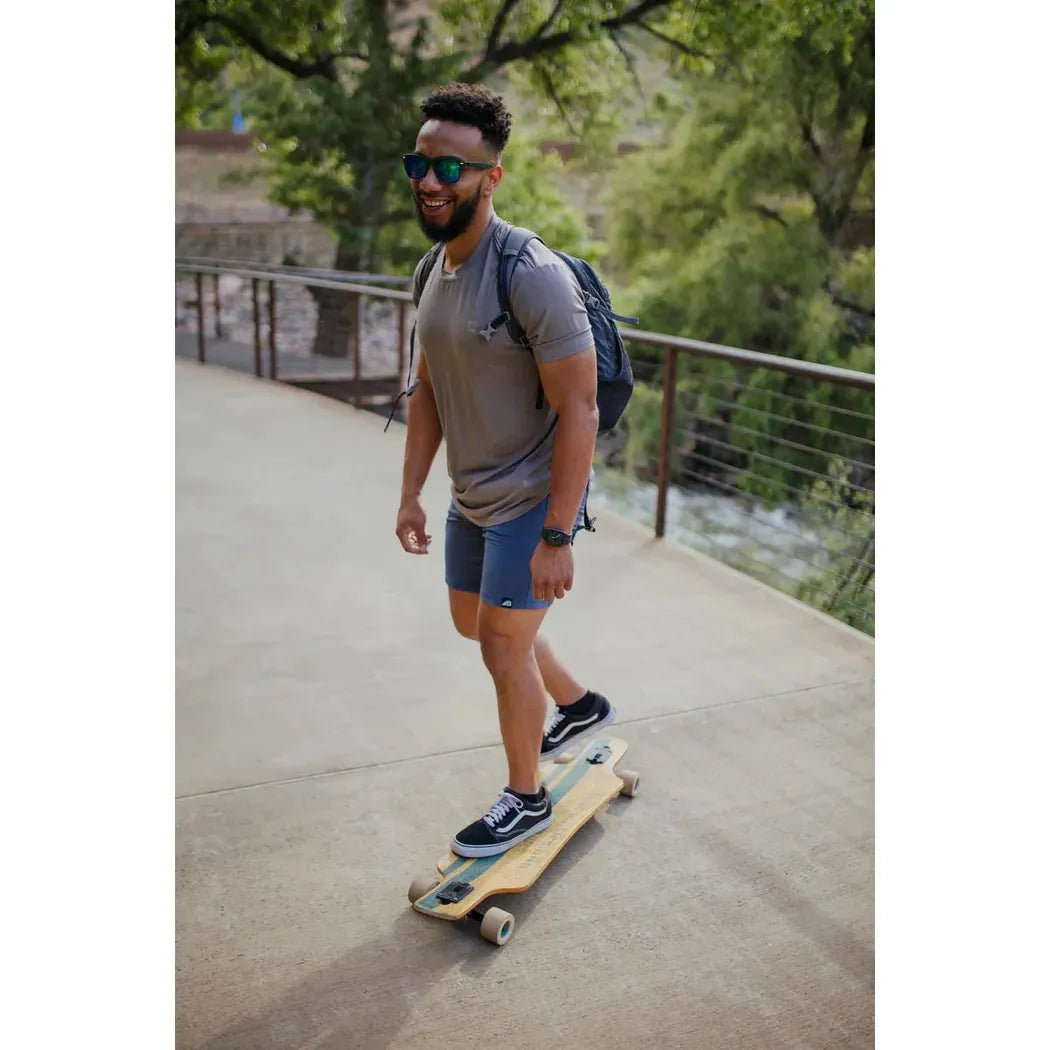 A man wearing the Belong Designs Men's Core Traverse Tek Tee in Granite rides a skateboard on a paved path with trees and a fence in the background.