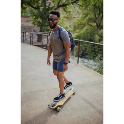 A man wearing the Belong Designs Men's Core Traverse Tek Tee in Granite rides a skateboard on a paved path with trees and a fence in the background.