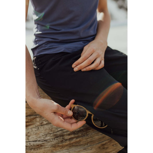 Close-up of a woman holding sunglasses while wearing Belong Designs Women's Rocky Mountain Joggers in Black, seated on a wooden surface outdoors.