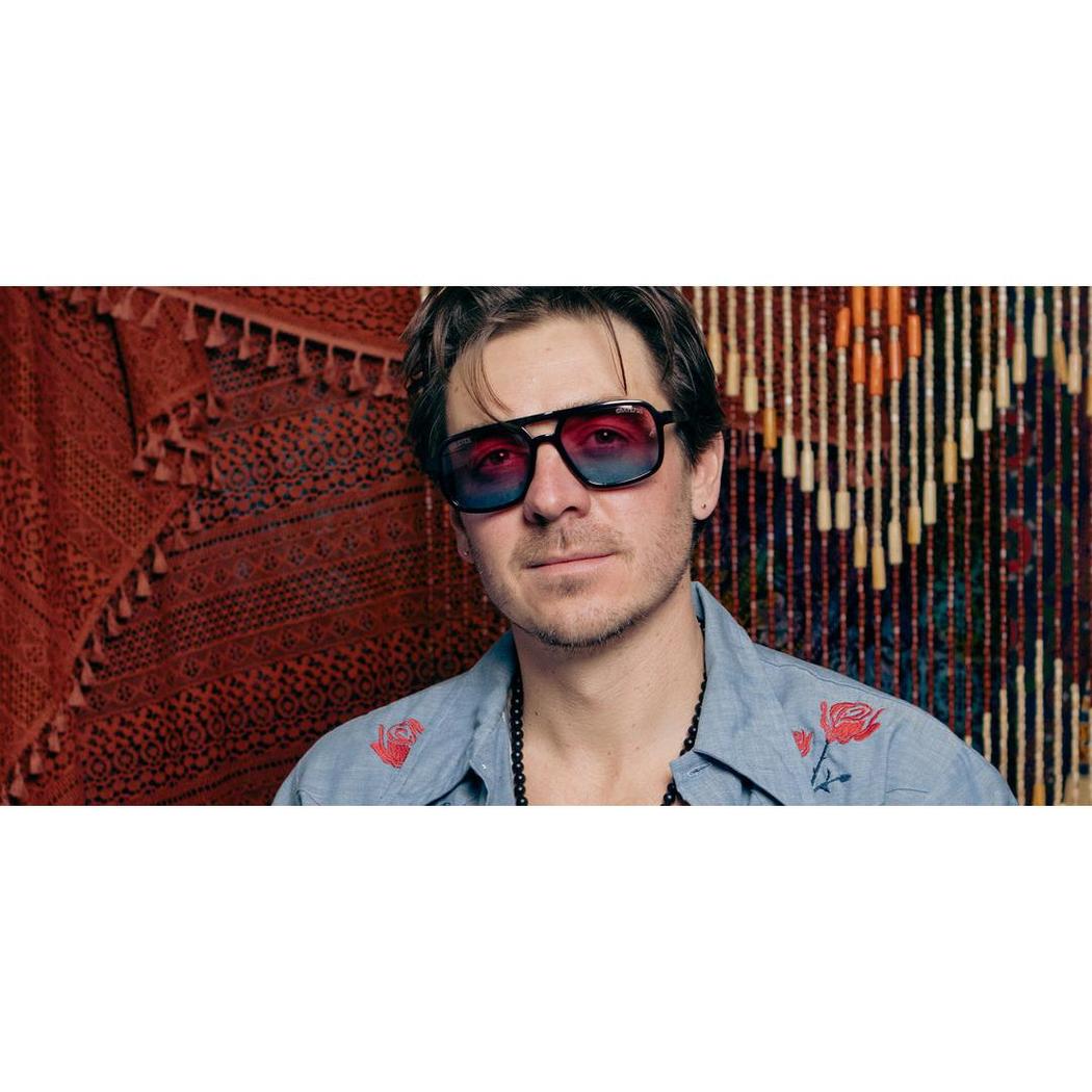 A model wears the Camp Eyewear Glacier: Grateful Dead Stealie Edition sunglasses with black frames and red lenses against a red and beaded background.