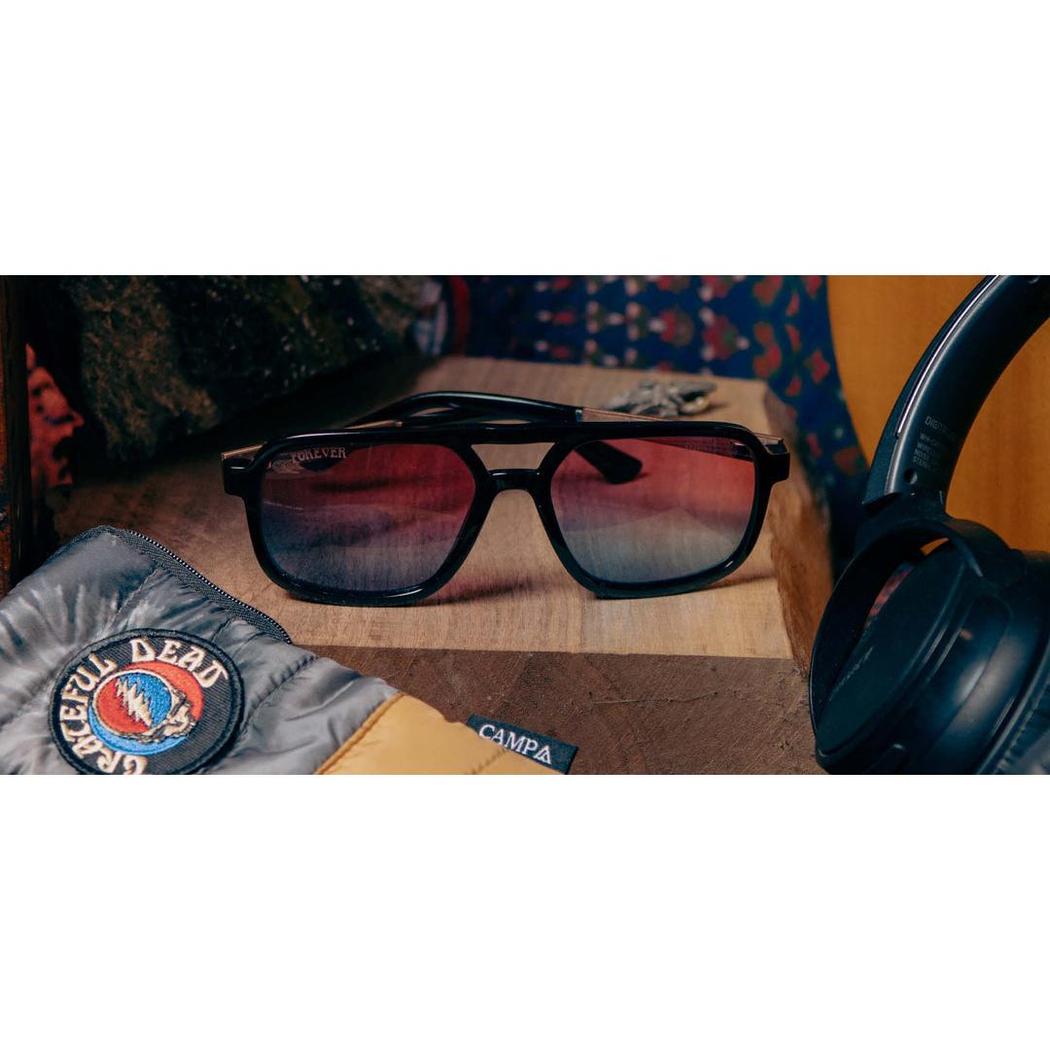 A close-up of black Camp Eyewear Glacier sunglasses with red-tinted lenses on a wooden surface, next to a Grateful Dead patch and headphones.