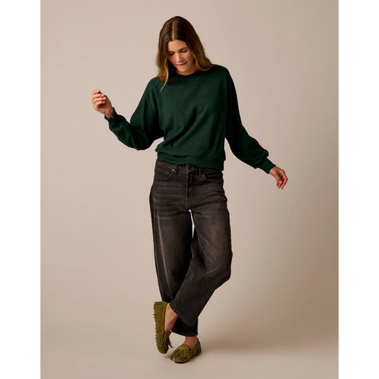 Model wearing the Carve Designs Anneke Supersoft Dolman in Dark Pine Heather with wide-leg jeans and green slippers against a neutral background.