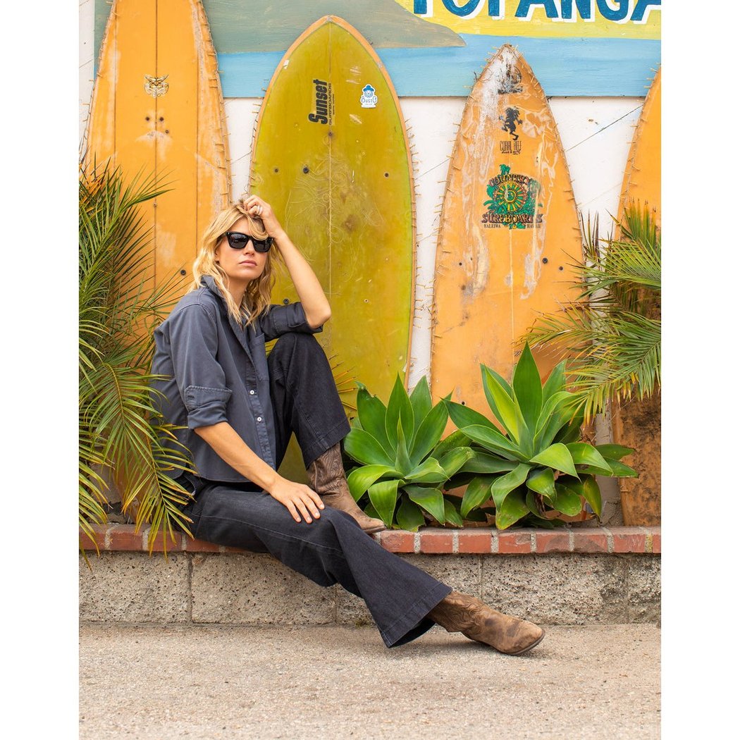 Woman in Washed Black Carve Designs Claire Twill Shirt sits on a ledge with surfboards and plants behind her.