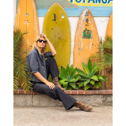 Woman in Washed Black Carve Designs Claire Twill Shirt sits on a ledge with surfboards and plants behind her.
