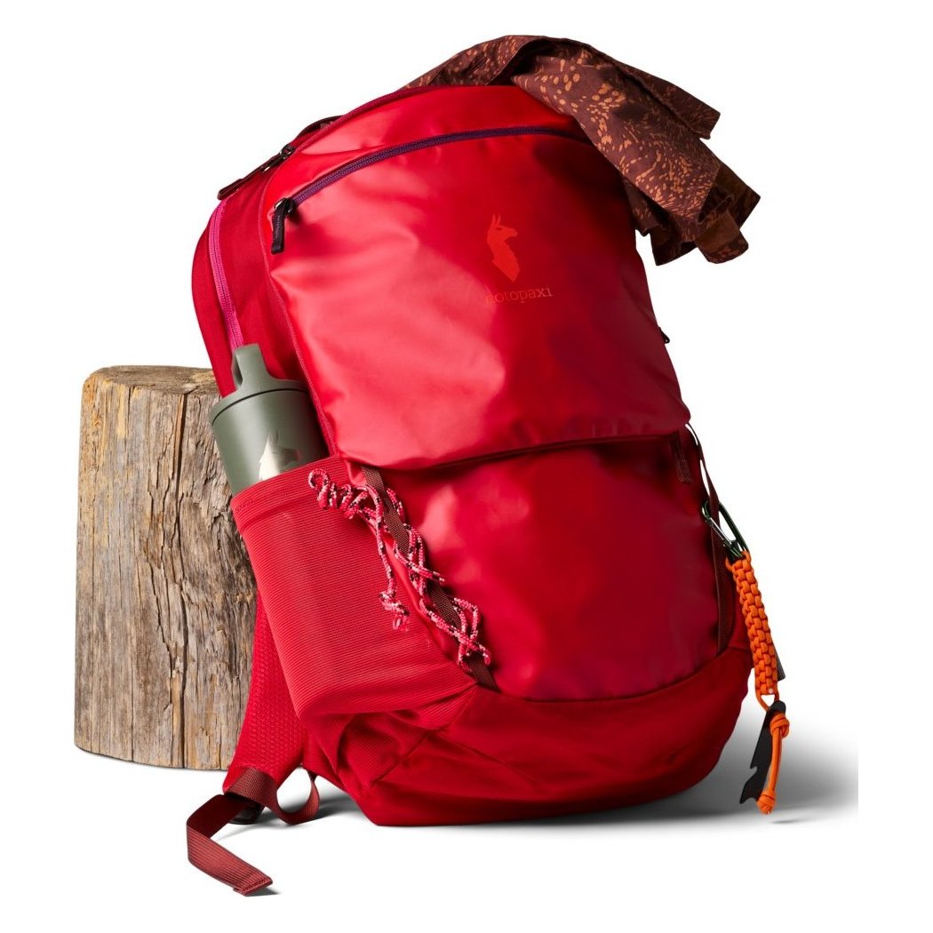 Front view of the Cotopaxi Allpa 26L Daypack in Blue Spruce/Abyss with a water bottle in the side pocket and a tree stump in the background.