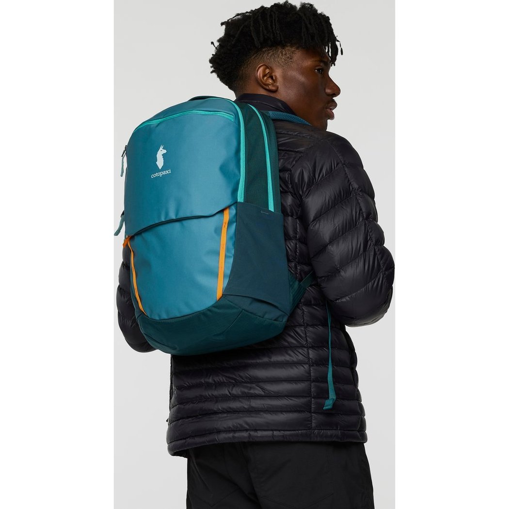 Back view of the Cotopaxi Allpa 26L Daypack in Blue Spruce/Abyss on a model wearing a black puffer jacket.