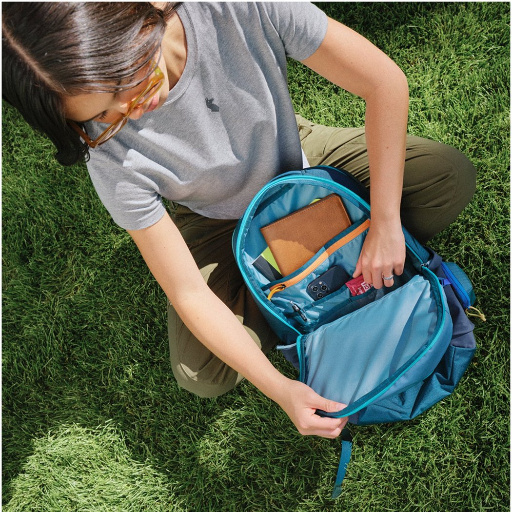 Overhead view of the Cotopaxi Allpa 26L Daypack in Blue Spruce/Abyss with a woman organizing items inside on green grass.