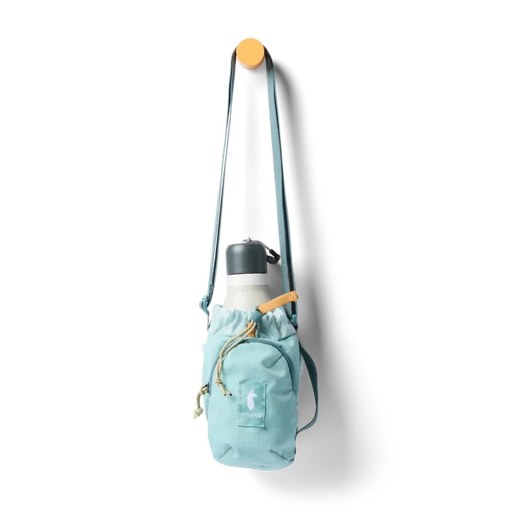 A light blue Cotopaxi Todo Water Bottle Sling hangs from a hook, holding a white water bottle with a charcoal lid, against a plain white background.