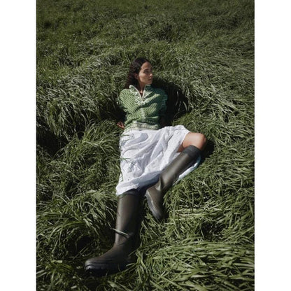 A woman wearing the Dale Of Norway Women's Leknes Sweater lies in tall green grass, showcasing the sweater and its offwhite skirt.