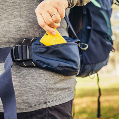 A person puts a yellow card into the pocket of a Gregory Amber 68 Plus backpack in Arctic Navy color, with a backpack strap visible and a grassy background.