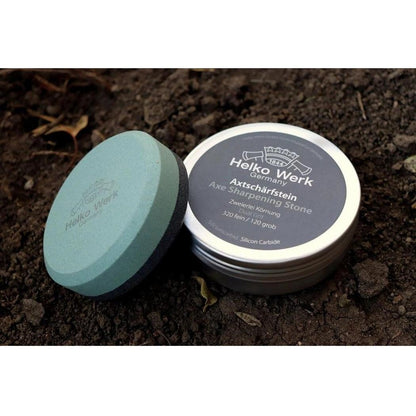 A round Helko Werk Axe Sharpening Stone in a tin sits on dark soil with its lid off.