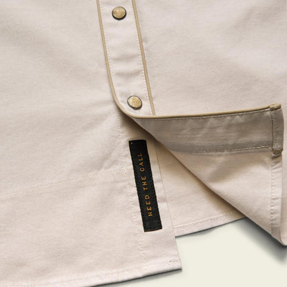 A close-up of the Howler Brothers Men's Broncbuster Snapshirt in Sand showing the snap placket and brand tag against a white background.