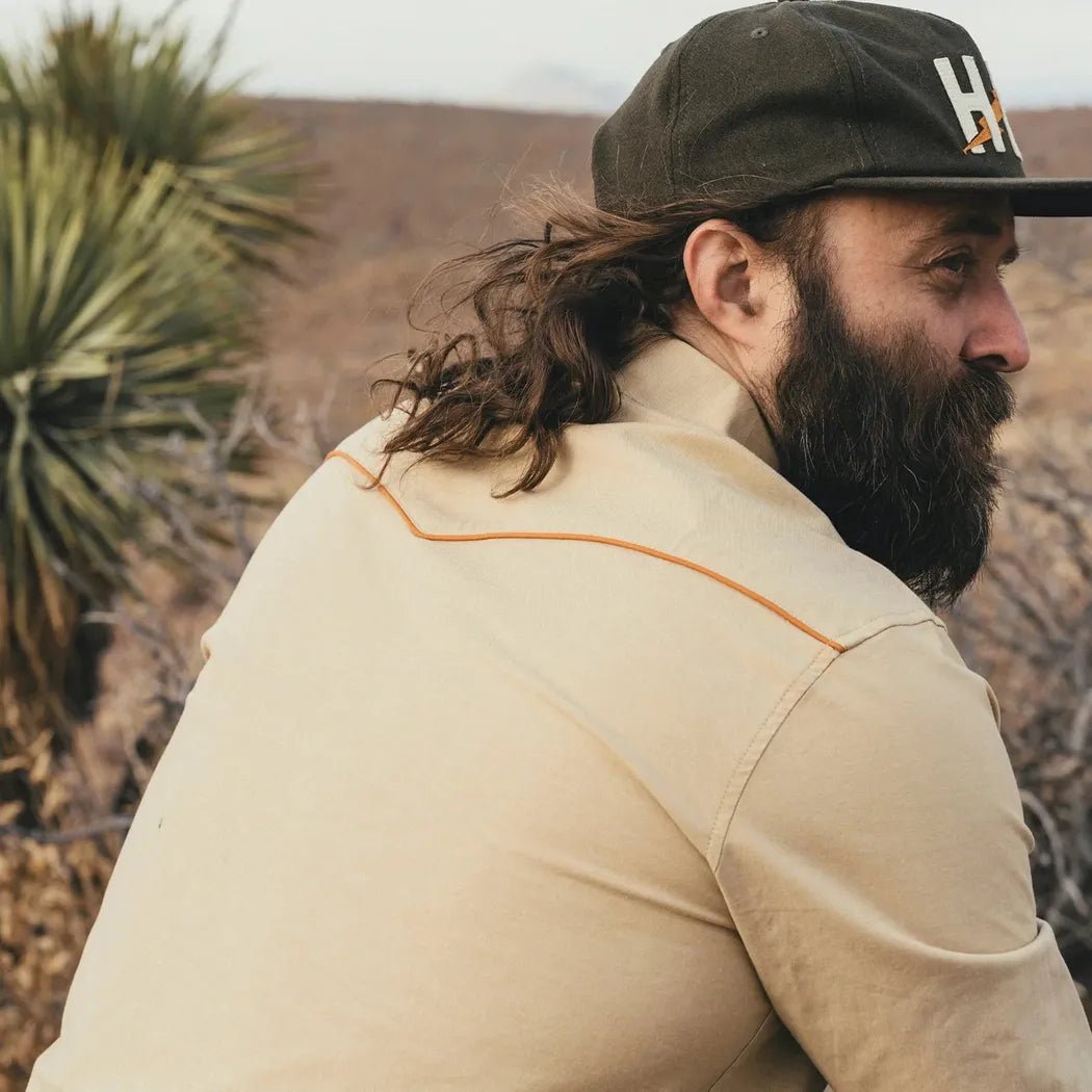 A man wearing the Howler Brothers Men's Crosscut Deluxe Longsleeve in El Sol : Gold poses outdoors with desert vegetation in the background.