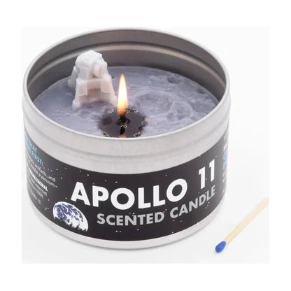 A close-up of the JD and Kate Industries Apollo 11 Scented Candle in a tin with a lit wick and a small astronaut figure, next to a matchstick.