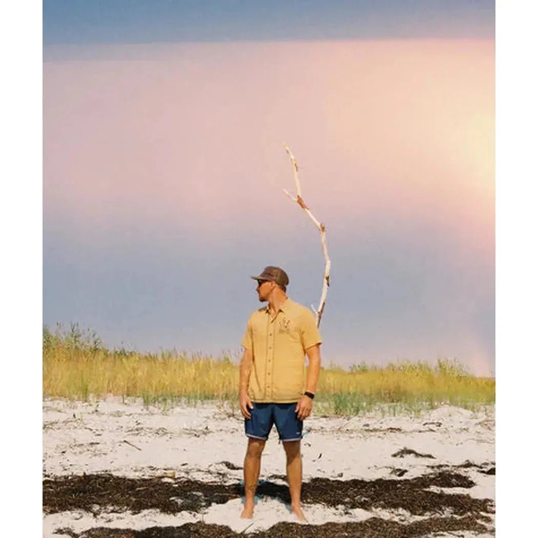 A man stands on a sandy beach wearing the Olive Jetty Men's Marshall Embroidered Linen Shirt, with a curved branch behind him and grassy dunes in the background.