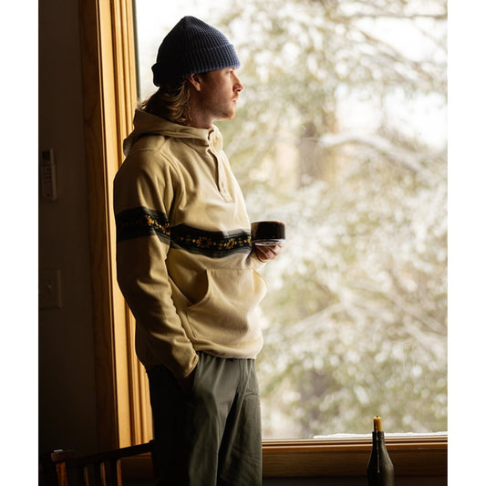 Man in Cream Jetty Quintin Fleece Hoodie stands by window with snow-covered trees outside, holding a mug.