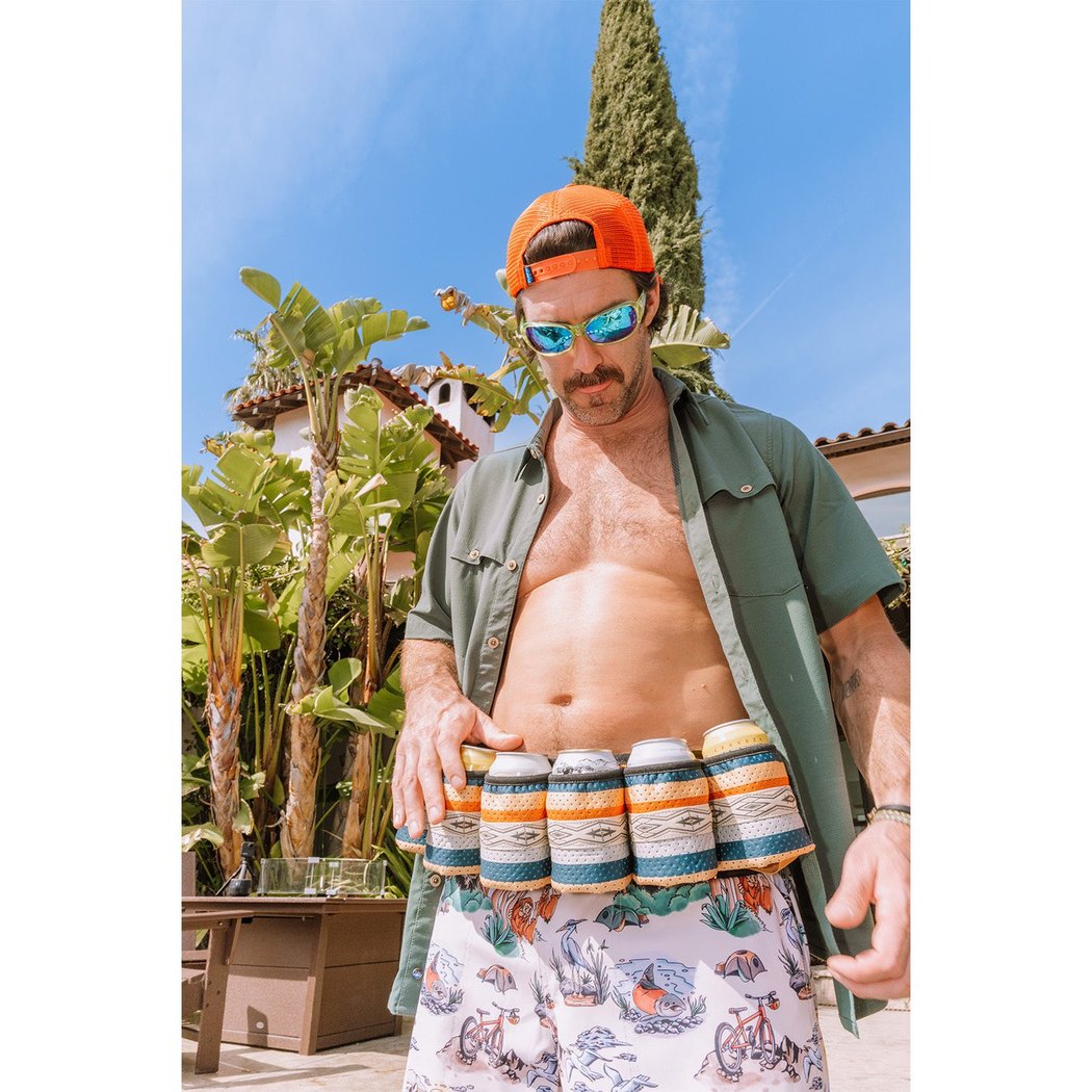 Man wearing the Kavu Beer Buddy in Park Lines with six cans, standing outdoors with palm trees and a blue sky.