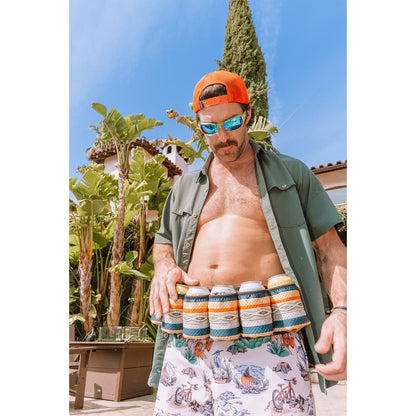 Man wearing the Kavu Beer Buddy in Park Lines with six cans, standing outdoors with palm trees and a blue sky.