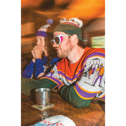 Two people wear Kavu Herschel accessories in Squirrely, one with a beanie and sunglasses, sitting at a wooden table with a metal cup.