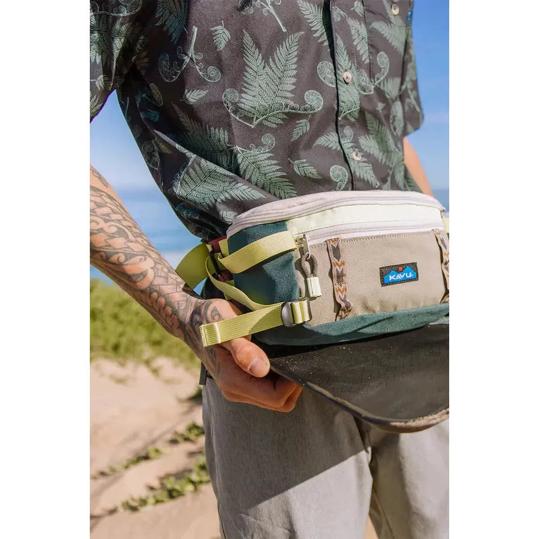 A man wearing a Fiddlehead Kavu Men's River Wrangler top holds the waist pack in front of a beach background.