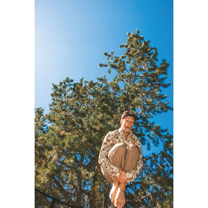 Model wearing the Kavu Westerly in Faraway Forest sits barefoot in a tree against a bright blue sky.