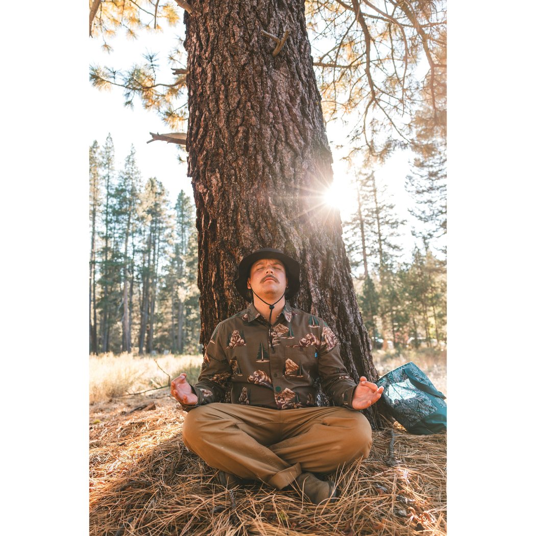 Man in Faraway Forest Kavu Westerly shirt meditating under a tree in a sunlit forest clearing.
