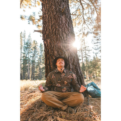 Man in Faraway Forest Kavu Westerly shirt meditating under a tree in a sunlit forest clearing.