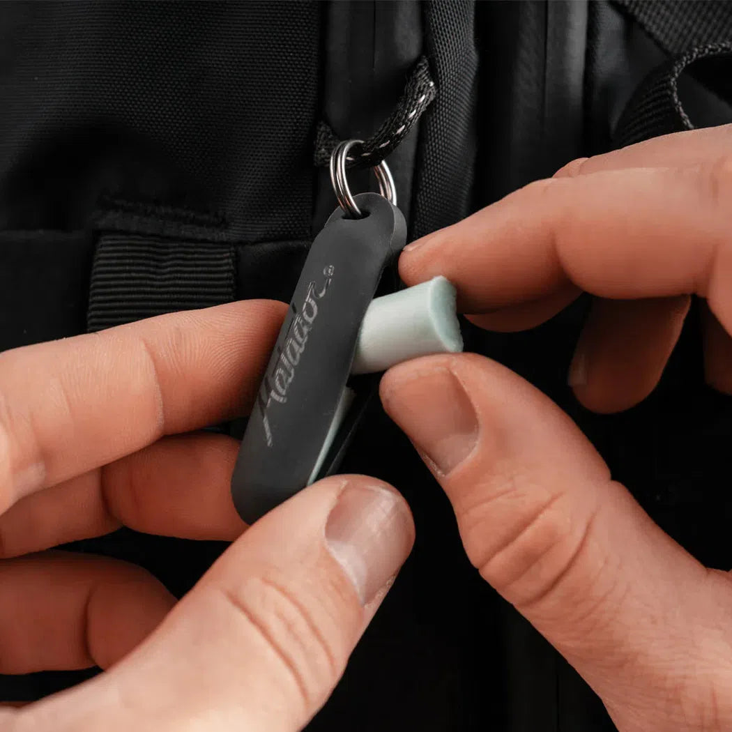 A person holding the Matador Travel Earplugs Kit, showing the charcoal-colored case and light green earplugs against a black backpack.