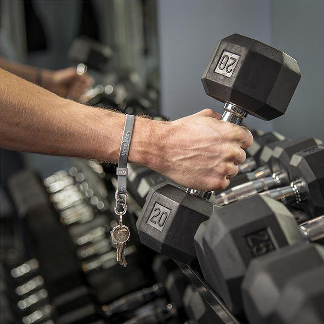 A hand wearing a blue Nite Ize Cinch-A-Lot Stretch Strap holds a 20-pound dumbbell in a gym setting.