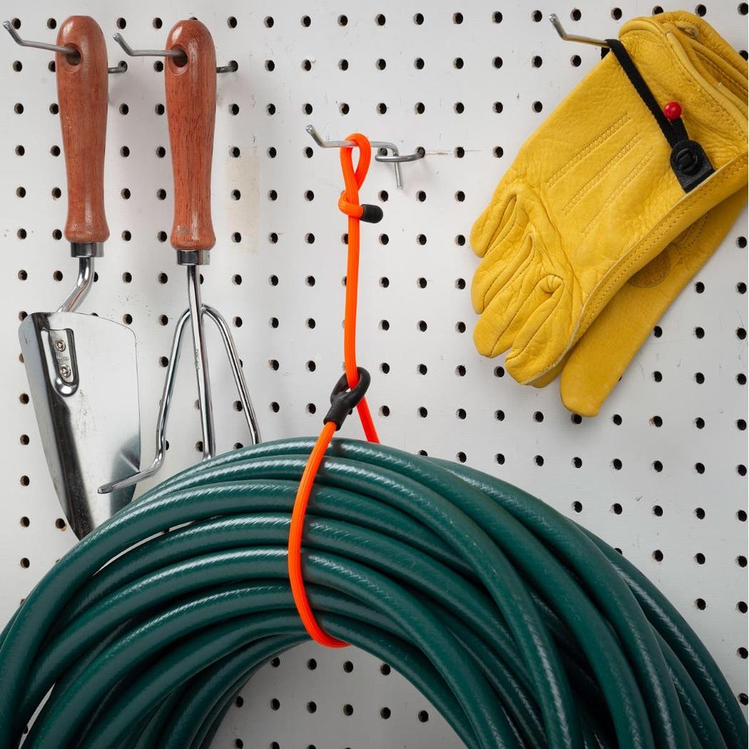 A bright orange Nite Ize Gear Tie Loopable Twist Tie hangs on a pegboard with gardening tools and yellow gloves in the background.