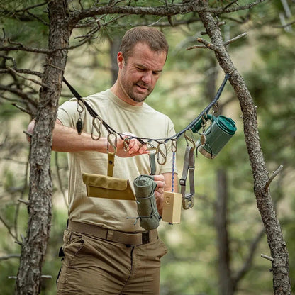 A man in a forest uses the Nite Ize GearLine Organization System with tactical gear hanging from tree branches.
