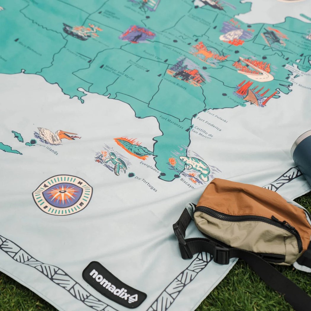 Nomadix Festival Blanket in Baja Aqua laid on grass with a brown bag and blue tumbler nearby.