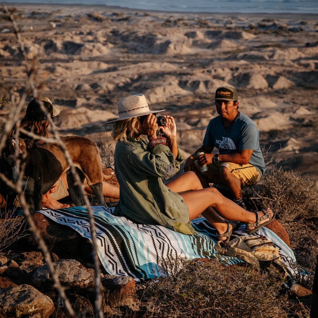 Woman in a hat sits on a Nomadix Festival Blanket in Baja Aqua while taking a photo with a desert landscape behind her.