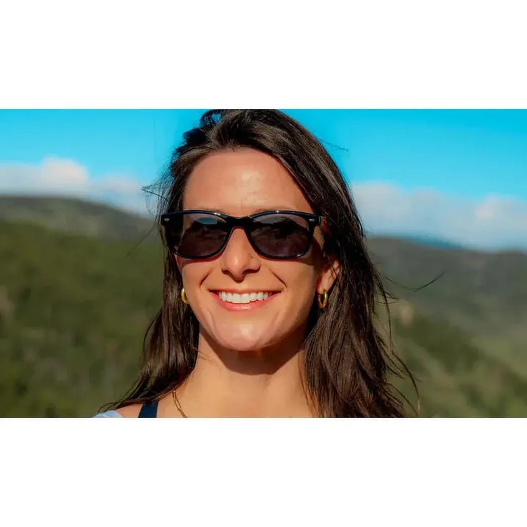 A woman smiles wearing the Shiny Black + Wood Optic Nerve Cruzin Shiny + Wood sunglasses against a mountain backdrop.