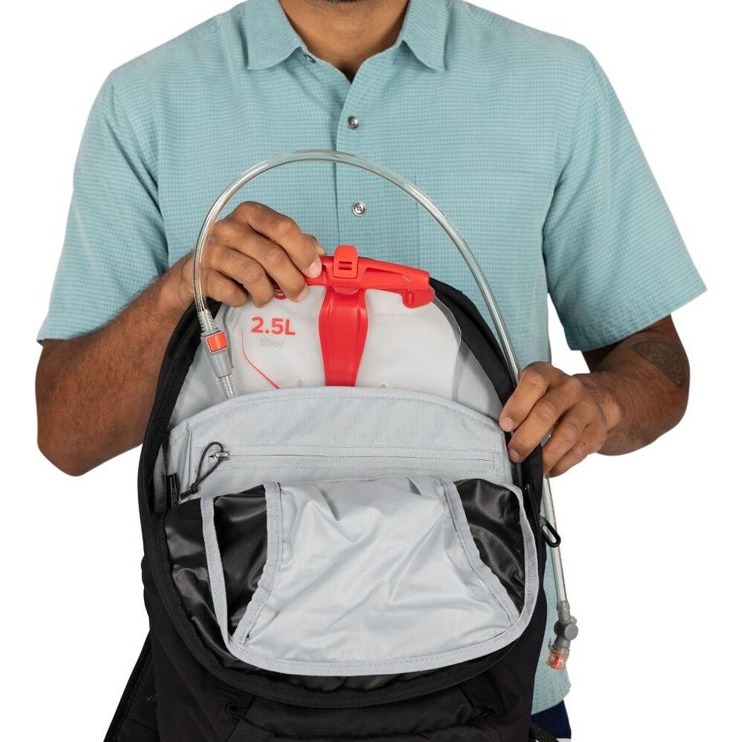 A person wearing a light blue shirt holds the Osprey Farpoint 55 black backpack open to show the 2.5L reservoir and hydration tube.