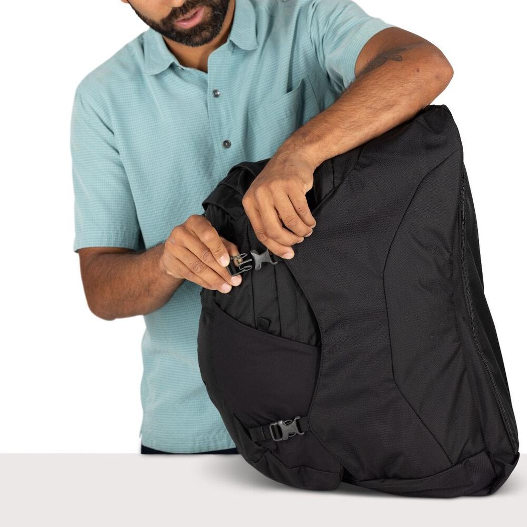 A man in a light blue shirt zips the black Osprey Farpoint 55 travel backpack on a white background.