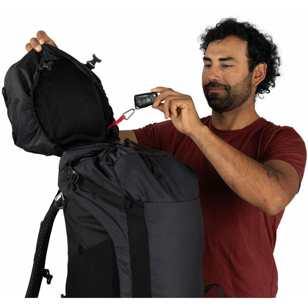A man in a red shirt holds a Moss Green Osprey Kestrel LT 65 backpack open, showing its top compartment and a car key attached to a red clip, against a plain white background.