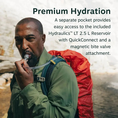 A man in a green shirt wears a Dark Charcoal Osprey Manta 34 Hiking Hydration Backpack with a red reservoir, drinking from a bite valve against a rocky background.
