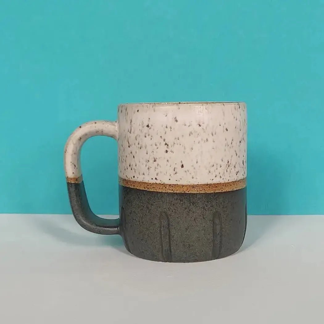 A side view of the Osso Ceramics Black and White Cookie Mug showing its speckled top and charcoal bottom against a turquoise background.