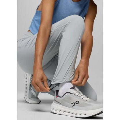 A woman crouching adjusts her grey blue Prana Women's Koen Mid Rise Daily Pant while wearing white On running shoes against a plain background.