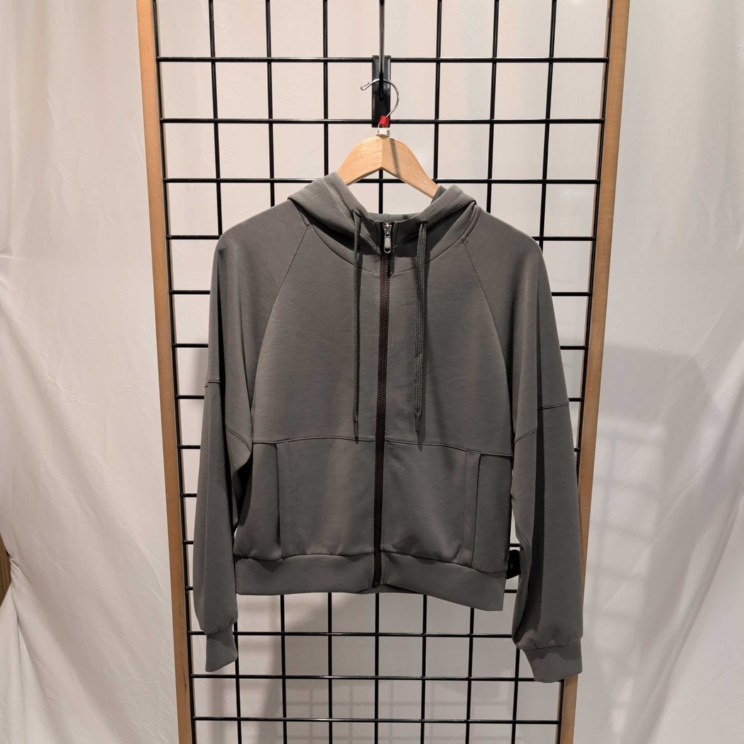 Front view of the Prana Womens Shae Full Zip-SAMPLE in Thyme hanging on a grid wall with a wooden hanger.